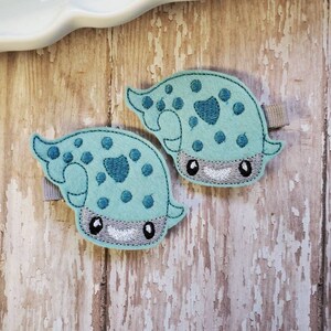 May include: Two teal blue felt appliques of a cartoon sea creature with black eyes, a white mouth, and gray accents. The appliques have a white felt backing and are attached to alligator clips.