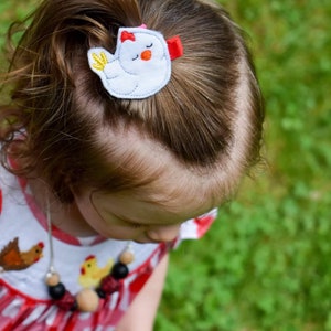 May include: A young girl with brown hair wearing a red and white gingham dress with a chicken design. She is wearing a necklace with wooden beads and a chicken hair clip with a red bow.