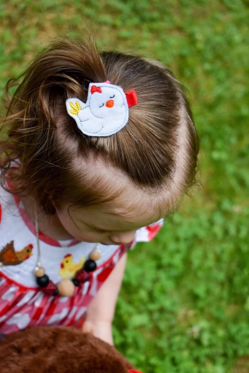 May include: A young girl with brown hair wearing a red and white gingham dress with a chicken design. She is wearing a necklace with wooden beads and a chicken hair clip with a red bow.