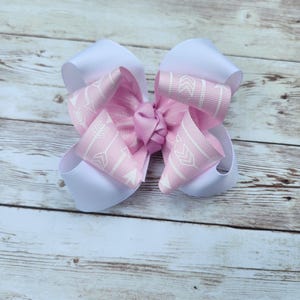 May include: A large, handmade hair bow featuring a layered design. The bow has a white base with a pink layer adorned with a white arrow pattern. The bow is tied in the center with a matching pink knot, perfect for accessorizing.