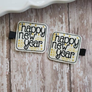 May include: Two square hair clips with the words "happy new year" embroidered in black on a white background. The design includes gold accents and a black clip on the back. The clips are on a weathered wooden surface.