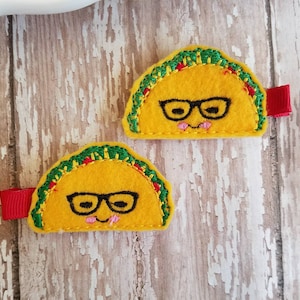 K&ouml;nnte beinhalten: Zwei gelbe Filz-Taco-Haarklammern mit gr&uuml;nem und rotem Besatz. Jeder Taco hat ein l&auml;chelndes Gesicht mit einer schwarzen Brille und einer rosa Zunge.