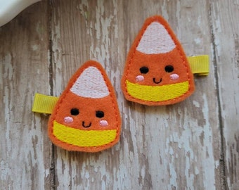 Puede incluir: Dos pinzas para el pelo de maíz dulce naranja, amarillo y blanco con caras sonrientes. Las pinzas tienen una base de cinta amarilla y están sobre un fondo de madera desgastada. El diseño de maíz dulce presenta ojos negros y mejillas rosas.