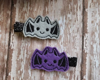 May include: Two felt bat hair clips. One is gray with black wings and a black smiling face. The other is purple with black wings and a black smiling face.