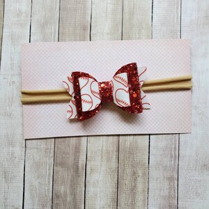 May include: Red and white glitter baseball bow headband. The bow is made of red glitter fabric with a white baseball print. The headband is made of a nude elastic.