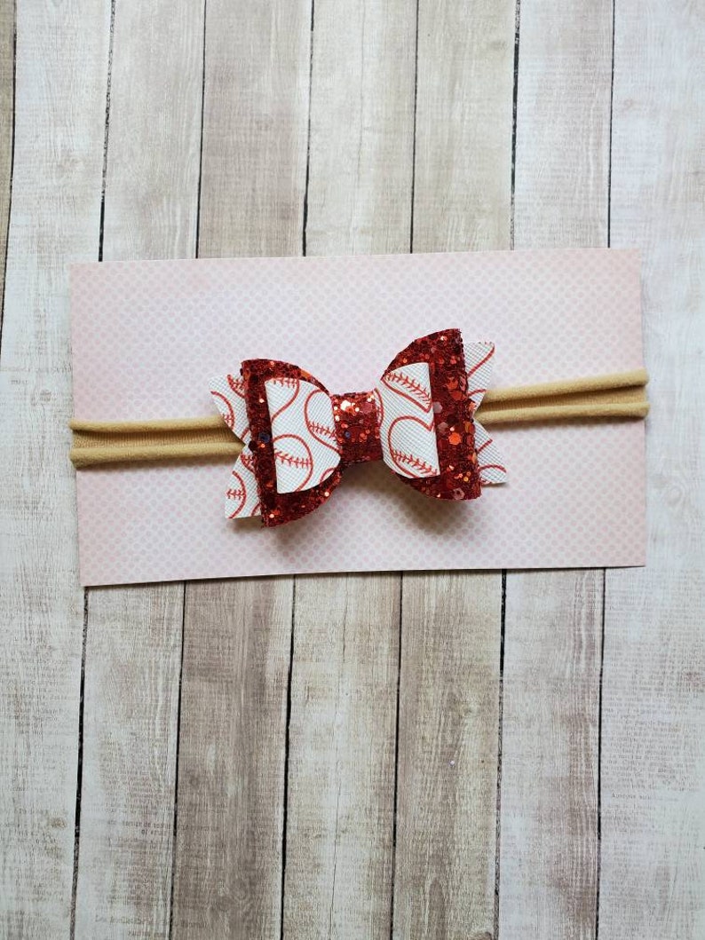 May include: Red and white glitter baseball bow headband. The bow is made of red glitter fabric with a white baseball print. The headband is made of a nude elastic.