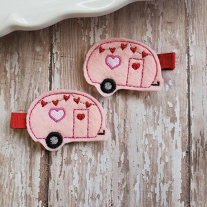 Puede incluir: Dos clips de camper de fieltro rosa con corazones de fieltro rojo y un corazón blanco en la parte delantera. Los clips tienen una cinta de fieltro roja en la parte posterior.