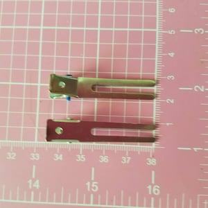 May include: Two silver hair clips with blue rubber tips. The clips are about 2 inches long and are shown on a pink grid background.
