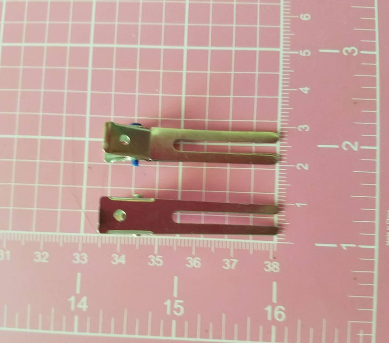 May include: Two silver hair clips with blue rubber tips. The clips are about 2 inches long and are shown on a pink grid background.
