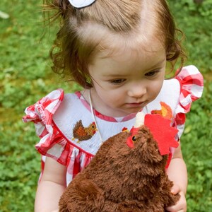 May include: A young child wearing a red and white gingham dress holds a brown plush chicken toy. The child has a white chicken hair clip in their hair.