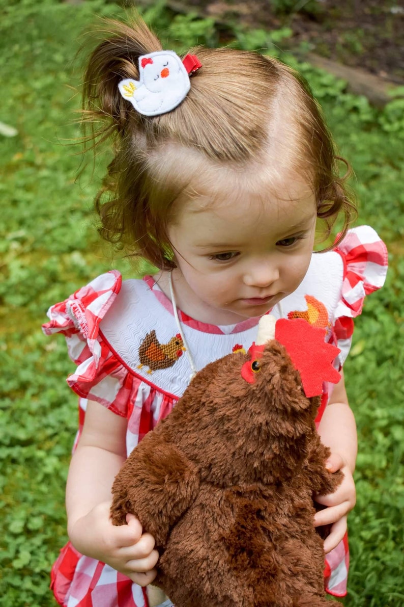 May include: A young child wearing a red and white gingham dress holds a brown plush chicken toy. The child has a white chicken hair clip in their hair.