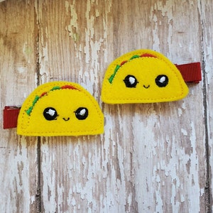 K&ouml;nnte beinhalten: Zwei gelbe Filz-Taco-Haarklammern mit gestickten l&auml;chelnden Gesichtern und gr&uuml;nen und roten Details.