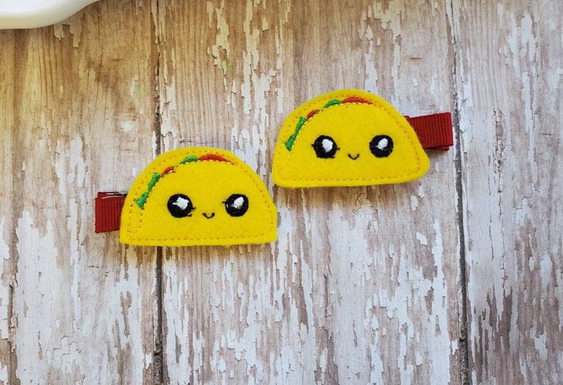 K&ouml;nnte beinhalten: Zwei gelbe Filz-Taco-Haarklammern mit gestickten l&auml;chelnden Gesichtern und gr&uuml;nen und roten Details.