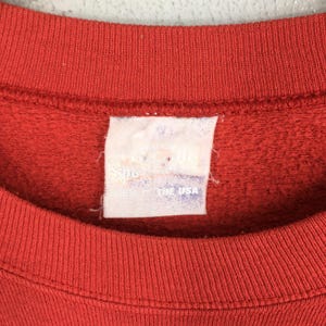 May include: Close-up of a red crew neck sweatshirt. The tag is visible, with text that is partially obscured. The tag reads "Made in the USA". The fabric has a textured appearance.
