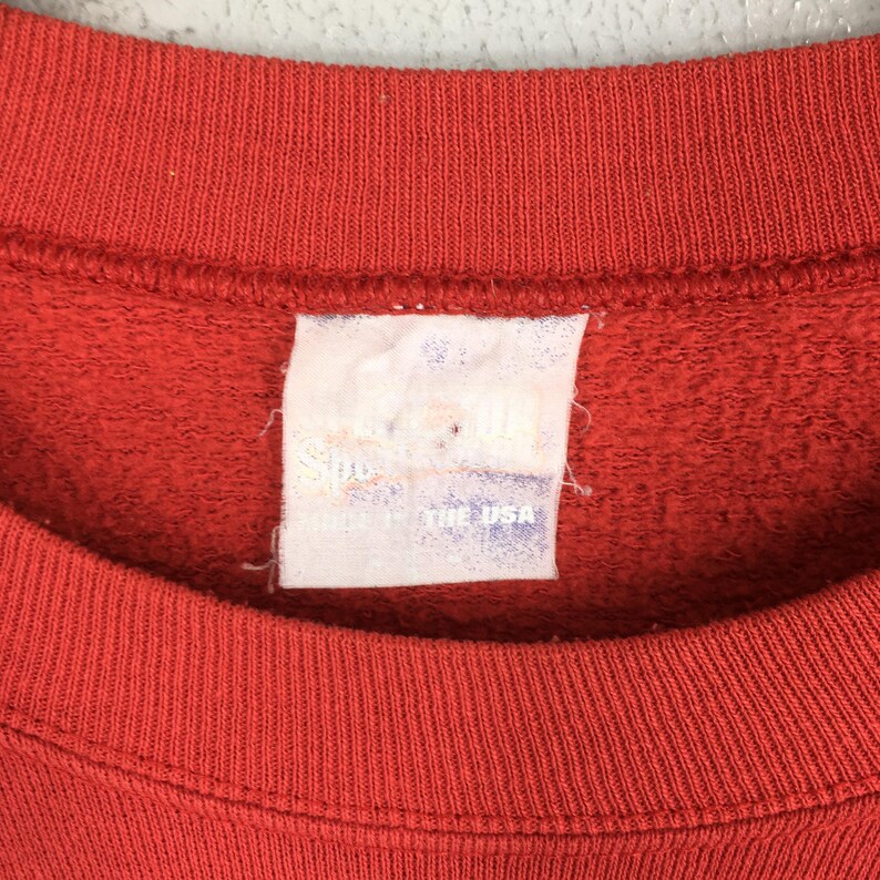 May include: Close-up of a red crew neck sweatshirt. The tag is visible, with text that is partially obscured. The tag reads "Made in the USA". The fabric has a textured appearance.