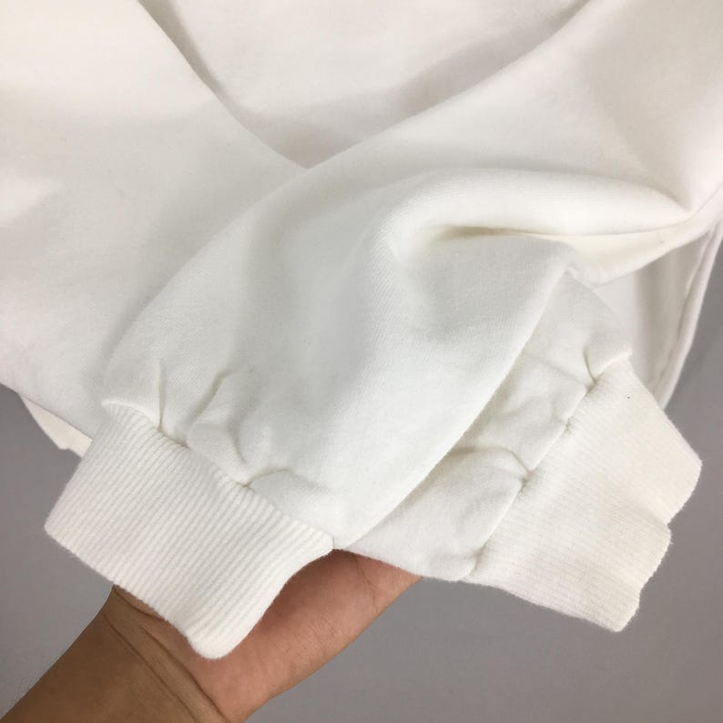Op de afbeelding: Close-up van een witte sweatshirtmouw. De manchet is geribbeld en de stof is bij de pols gerimpeld. De mouw is gemaakt van een zacht, dik materiaal.