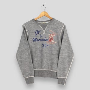 May include: A heather gray sweatshirt with white stitching and a crew neck. The front features the text "Joe The Marauder 22nd" in blue and a red graphic of a winged figure. The sweatshirt is hanging on a wooden hanger.