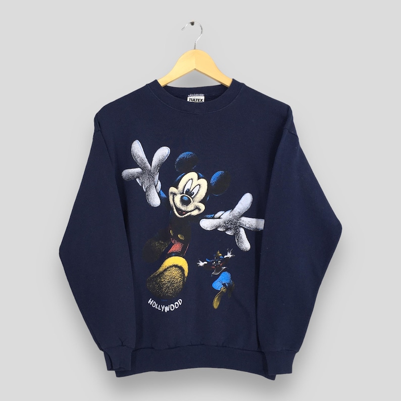 May include: Navy blue sweatshirt featuring a large graphic of Mickey Mouse with outstretched arms and a smaller image of Minnie Mouse. The word "Hollywood" is printed in white. The sweatshirt is hanging on a wooden hanger.