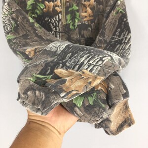 K&ouml;nnte beinhalten: Ein Sweatshirt mit Camouflage-Muster und einem Viertelrei&szlig;verschluss. Der Stoff weist eine Mischung aus braunen, gr&uuml;nen und grauen Farbt&ouml;nen auf, die eine Waldumgebung nachahmen. Die B&uuml;ndchen und der Kragen sind gerippt.