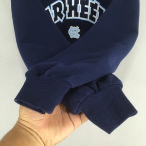 May include: Navy blue sweatshirt with the word "RHEE" in white block letters and a light blue emblem. The cuffs are ribbed and the sleeves are folded. The sweatshirt is made of a soft, comfortable material.