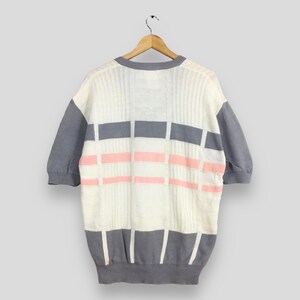 May include: A short-sleeved sweater in a cream color with gray sleeves and trim. The front features a geometric pattern of gray and pink rectangles. The sweater is hanging on a wooden hanger against a light gray background.
