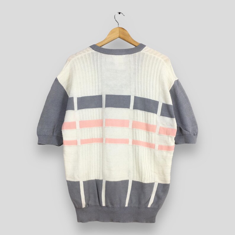 May include: A short-sleeved sweater in a cream color with gray sleeves and trim. The front features a geometric pattern of gray and pink rectangles. The sweater is hanging on a wooden hanger against a light gray background.