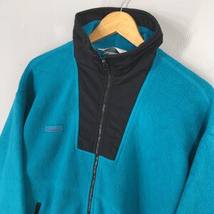 May include: A teal fleece jacket with a black panel on the chest and collar. The jacket has a full-length zipper and a small logo patch on the left chest. The collar is a high, stand-up style.