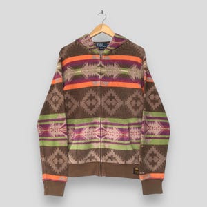May include: A brown zip-up hoodie featuring a multi-colored geometric pattern in orange, purple, and light green. The garment includes a hood, long sleeves with ribbed cuffs, and a Polo by Ralph Lauren label at the neckline and lower hem.