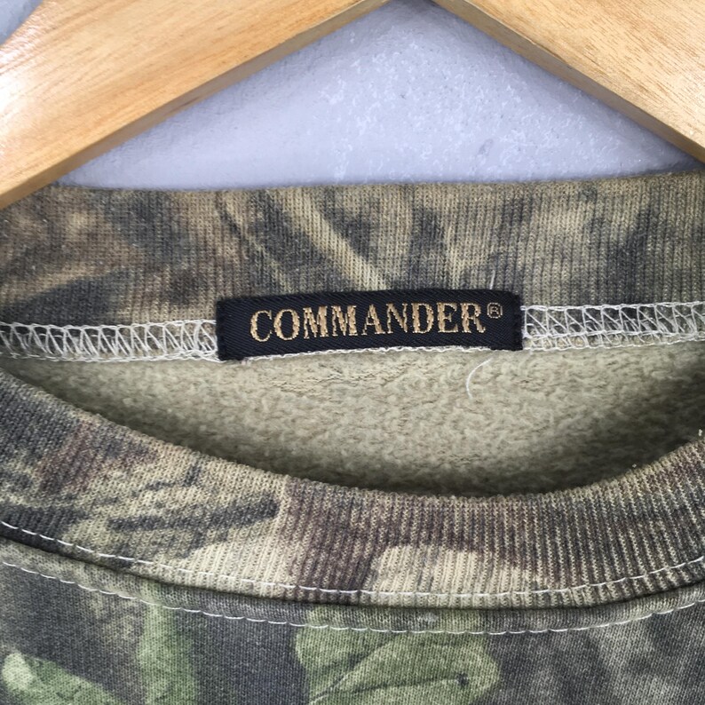 K&ouml;nnte beinhalten: Ein Sweatshirt mit Camouflage-Muster und dem Wort "COMMANDER" auf einem schwarzen Etikett. Das Sweatshirt hat einen Rundhalsausschnitt und ein strukturiertes, gesprenkeltes Design in Gr&uuml;n-, Braun- und Beiget&ouml;nen.