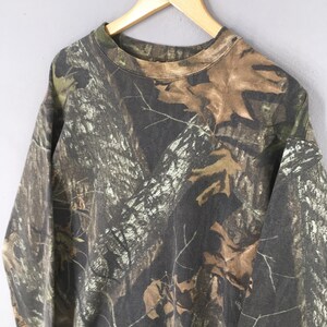 Puede incluir: Una sudadera de camuflaje de manga larga cuelga de una percha de madera. El patr&oacute;n presenta tonos de marr&oacute;n, verde y negro, que se asemejan a una escena forestal con hojas y corteza de &aacute;rbol. Se ven el cuello redondo y las mangas.