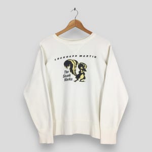 May include: Off-white crewneck sweatshirt with the text "LOCKHEED MARTIN" above a cartoon skunk graphic. The skunk is black and yellow, with the text "The Skunk Works" below it. The sweatshirt is hanging on a wooden hanger.