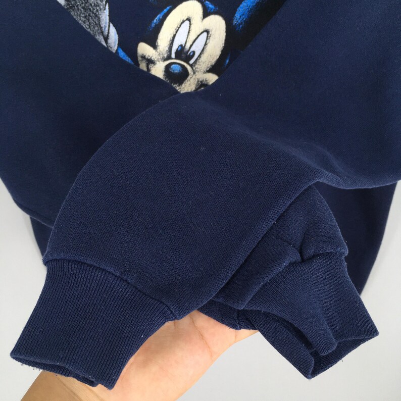 May include: Navy blue sweatshirt featuring a Mickey Mouse graphic. The graphic shows Mickey Mouse's face in white, black, and blue. The sweatshirt has ribbed cuffs and a crew neck. The fabric appears to be a soft, comfortable material.