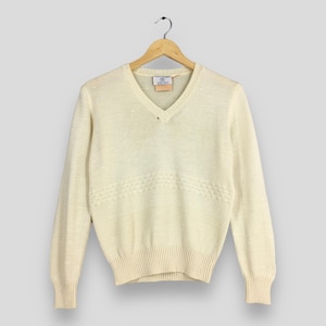 May include: Cream-colored V-neck sweater with long sleeves, hanging on a wooden hanger. The sweater features a textured knit design with a decorative horizontal pattern across the chest. A small label is visible near the neckline.
