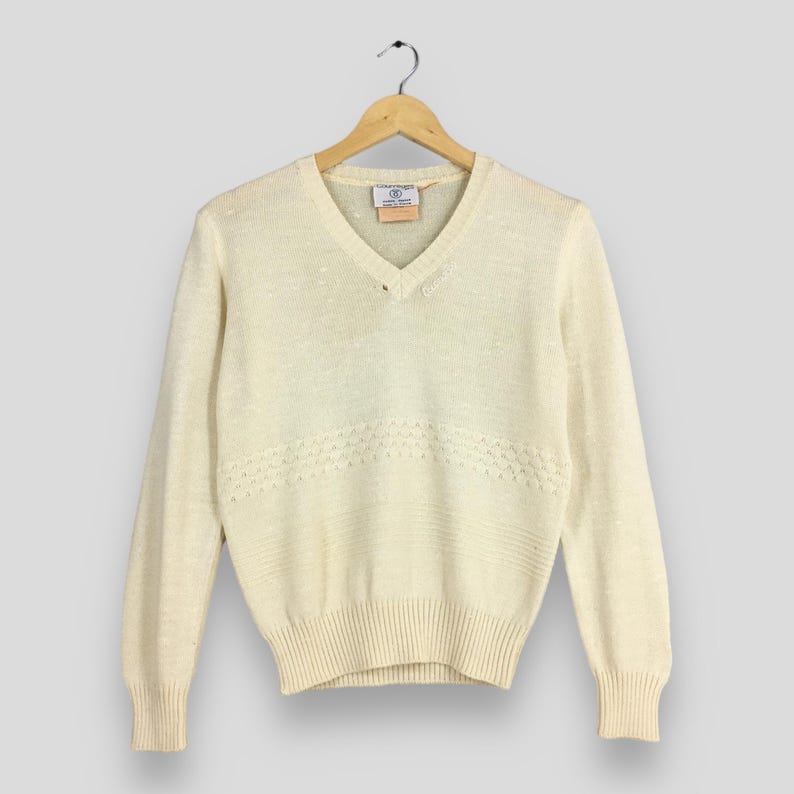 May include: Cream-colored V-neck sweater with long sleeves, hanging on a wooden hanger. The sweater features a textured knit design with a decorative horizontal pattern across the chest. A small label is visible near the neckline.