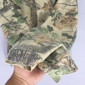 Puede incluir: Primer plano de una manga de sudadera con estampado de camuflaje. La tela presenta una mezcla de tonos verdes, marrones y beige, imitando un entorno forestal. El pu&ntilde;o es de color beige liso.