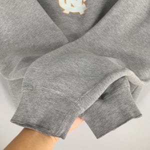May include: A gray sweatshirt with a light blue and gold emblem on the chest. The sweatshirt has ribbed cuffs and a crew neck. The fabric appears to be a soft, comfortable material, suitable for casual wear.