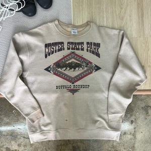 May include: A beige crewneck sweatshirt featuring a graphic print with the text CUSTER STATE PARK and BUFFALO ROUNDUP. The design includes a central diamond shape with a geometric pattern and an illustration of buffalo being herded by a rider.