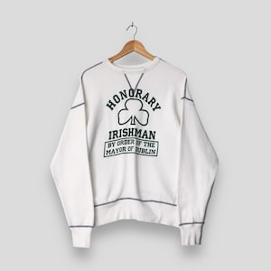 以下が含まれることがあります： クリーム色のスウェットシャツ。緑色の「HONORARY IRISHMAN」の文字とクローバーのグラフィック、そして「BY ORDER OF THE MAYOR OF DUBLIN」の文字がプリントされています。濃いグレーのステッチが施されています。