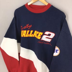 May include: Navy blue sweatshirt with red and white accents. The front features the name "Rusty Wallace" in red and yellow, along with the number "2" and a signature. A circular patch with the word "Miller" is on the sleeve.