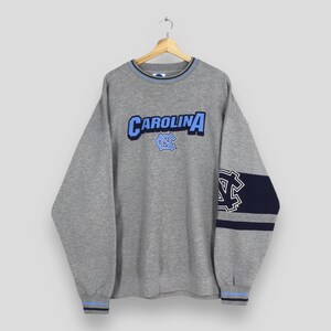 Puede incluir: Una sudadera gris con la palabra "CAROLINA" en letras azules y blancas en el pecho. La sudadera tiene detalles en azul y marino, incluyendo un logotipo en la manga y alrededor del cuello y los pu&ntilde;os. Cuelga de una percha de madera.