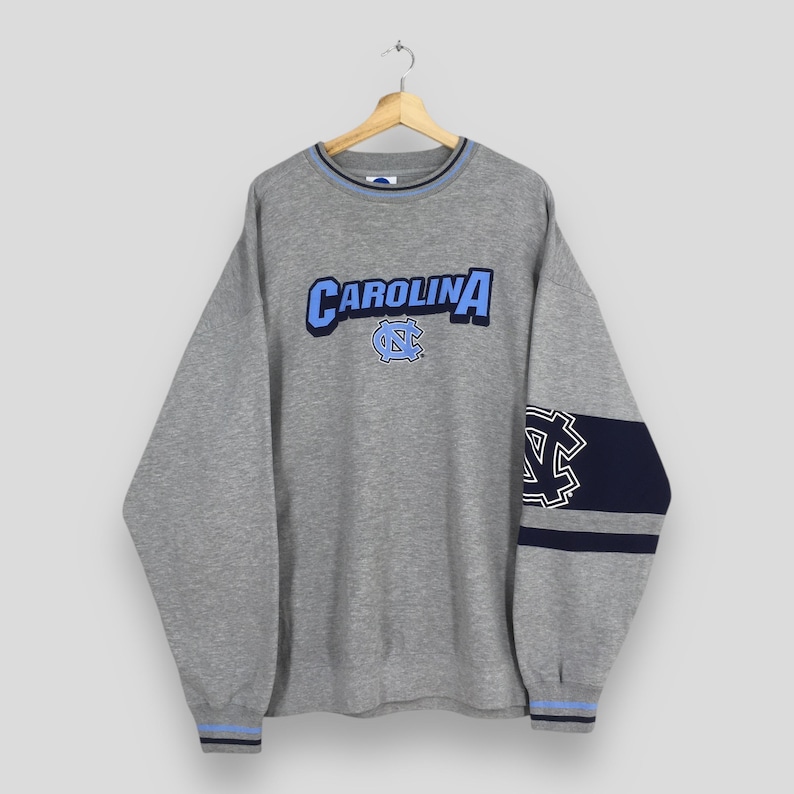 Puede incluir: Una sudadera gris con la palabra "CAROLINA" en letras azules y blancas en el pecho. La sudadera tiene detalles en azul y marino, incluyendo un logotipo en la manga y alrededor del cuello y los pu&ntilde;os. Cuelga de una percha de madera.