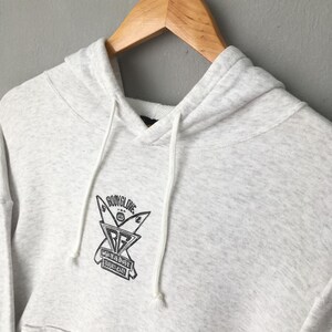 Puede incluir: Una sudadera con capucha gris claro con capucha con cord&oacute;n y bolsillo de canguro. La sudadera con capucha presenta un gr&aacute;fico negro en el pecho con el texto "BODYGLOVE" y "WHEN IN DOUBT PADDLE OUT". La sudadera con capucha est&aacute; colgada en una percha de madera.