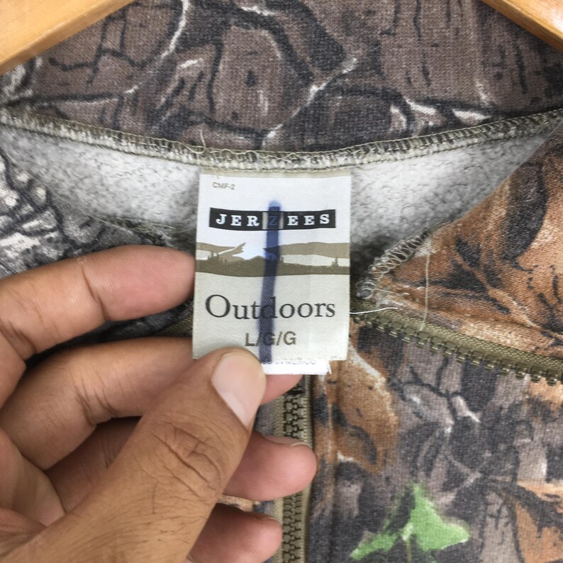 K&ouml;nnte beinhalten: Ein Sweatshirt mit Camouflage-Muster, Rei&szlig;verschluss und einem Etikett mit der Aufschrift "JERZEES Outdoors L/G". Das Sweatshirt weist eine Mischung aus braunen, gr&uuml;nen und grauen Farben auf, die sich in die Au&szlig;enumgebung einf&uuml;gen sollen.