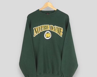 Może przedstawiać: Ciemnozielona bluza z napisem "NOTRE DAME" w kolorze żółtym, wygiętym na piersi. Poniżej okrągła odznaka z literami "ND". Bluza wisi na drewnianym wieszaku.