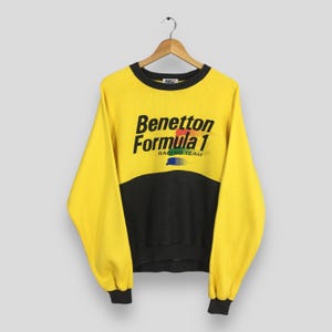 Puede incluir: Una sudadera amarilla y negra de Benetton Formula 1 Racing Team. La sudadera tiene cuello, puños y dobladillo negros. El logotipo de Benetton Formula 1 está impreso en negro con un acento arcoíris. La sudadera está colgada de una percha de madera.