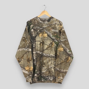 May include: A long-sleeved camouflage sweatshirt with a crew neck, hanging from a wooden hanger. The pattern features a mix of brown, green, and tan, resembling a forest scene. The sweatshirt has a small logo on the upper right chest.