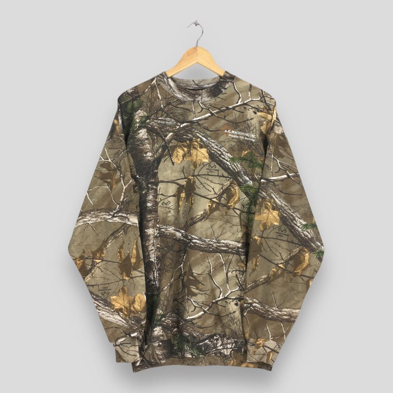 May include: A long-sleeved camouflage sweatshirt with a crew neck, hanging from a wooden hanger. The pattern features a mix of brown, green, and tan, resembling a forest scene. The sweatshirt has a small logo on the upper right chest.
