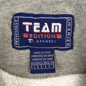 May include: Close-up of a navy blue tag with white text that reads "TEAM EDITION APPAREL". The tag also includes the text "OFFICIALLY LICENSED" and "MADE IN CHINA". The tag is on a gray fabric.