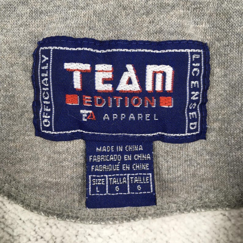 May include: Close-up of a navy blue tag with white text that reads "TEAM EDITION APPAREL". The tag also includes the text "OFFICIALLY LICENSED" and "MADE IN CHINA". The tag is on a gray fabric.