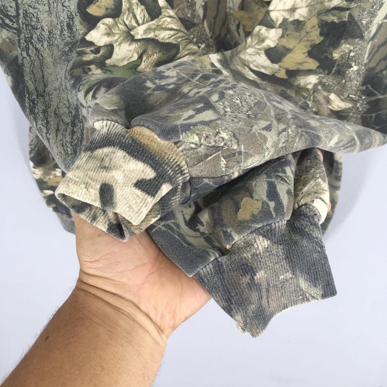 May include: A camouflage-patterned sweatshirt in shades of green, brown, and gray. The cuffs and collar are ribbed, with a faded appearance. The fabric has a detailed leaf and branch design, typical of hunting apparel.